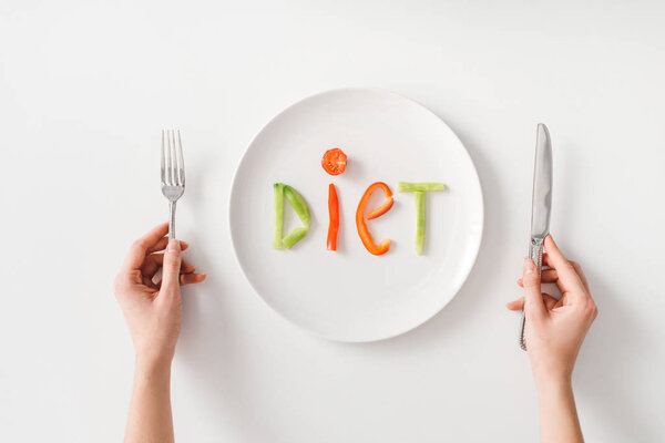 Top view of woman holding fork and knife near lettering diet from vegetable slices on plate on white background
