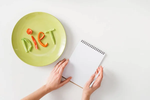 Top view of woman with pen and notebook near plate with diet lettering from vegetable slices on white background