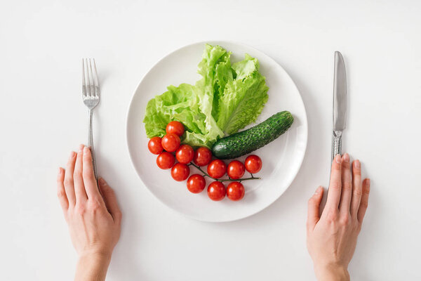 Top view of woman holding cutlery near fresh vegetables on plate on white background