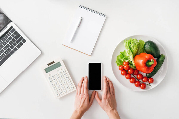 Top view of woman holding smartphone near calculator, notebook and vegetables on white background