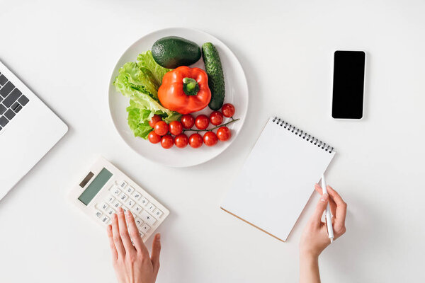 Top view of woman using calculator near digital devices, notebook and fresh vegetables on white background
