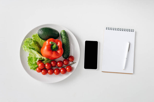 Top view of vegetables and avocado on plate, smartphone and notebook on white background