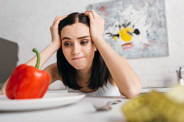 Selective focus of pensive woman looking at bell pepper near measuring tape on table