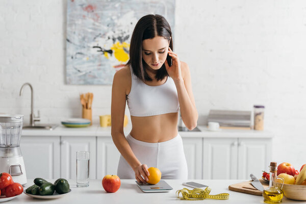 Beautiful sportswoman weighing orange with scales near measuring tape and smartphone on kitchen table, calorie counting diet