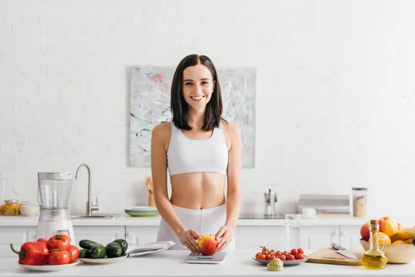 Smiling Sportswoman Weighing Fresh Apple Measuring Tape Notebook Kitchen Table — Stockfoto