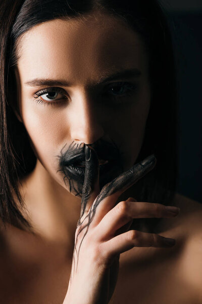 Beautiful nude woman with black painted hand and lips showing silence gesture and looking at camera isolated on black 