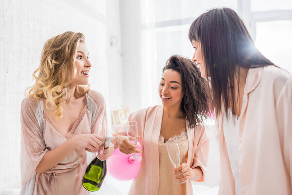 счастливые мультикультурные девушки открывают бутылку шампанского на девичнике
