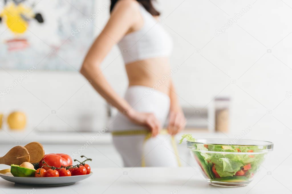 Selective focus of organic vegetables with salad on table and sportswoman measuring hips with tape in kitchen