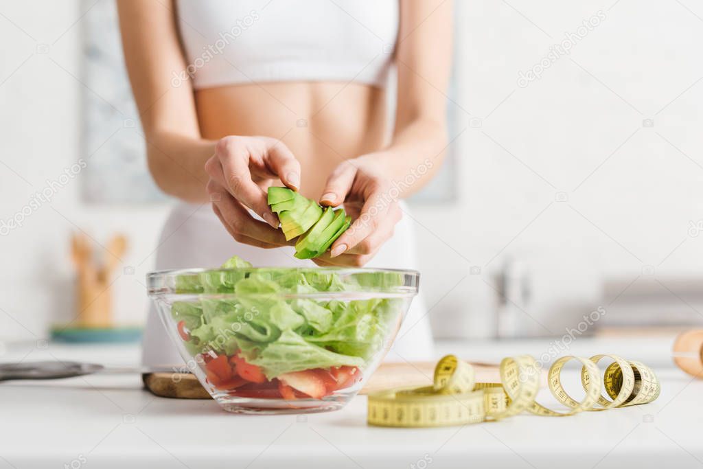 Cropped view of slim woman cooking salad with fresh vegetables and avocado near measuring tape on kitchen table