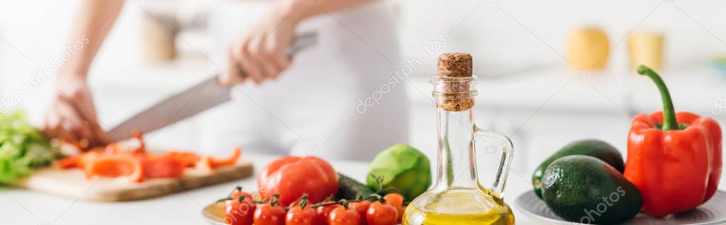 Selective focus of olive oil and fresh vegetables near woman cooking salad on kitchen table, panoramic shot