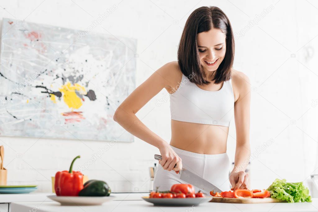 Smiling sportswoman cutting fresh vegetables and lettuce on kitchen table