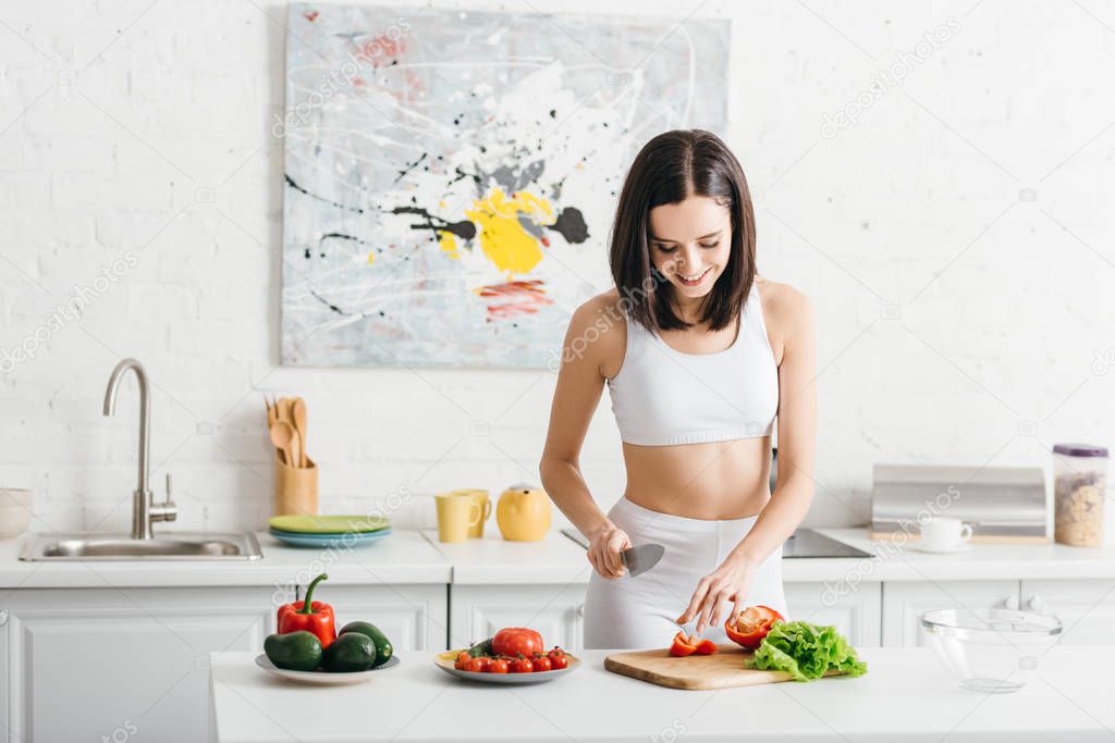 Smiling sportswoman cooking salad with ripe vegetables on kitchen table