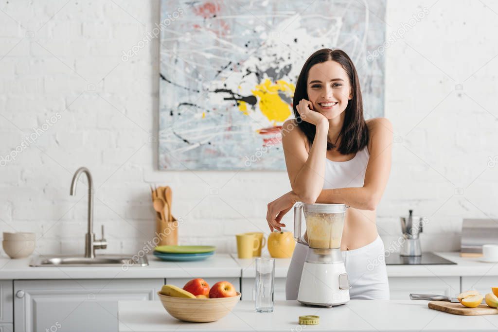 Slim sportswoman smiling at camera while preparing smoothie near measuring tape on kitchen table