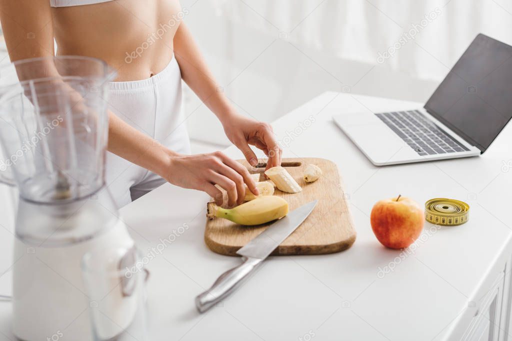 Cropped view of slim girl in sportswear cutting fruits for smoothie near laptop and measuring tape on table