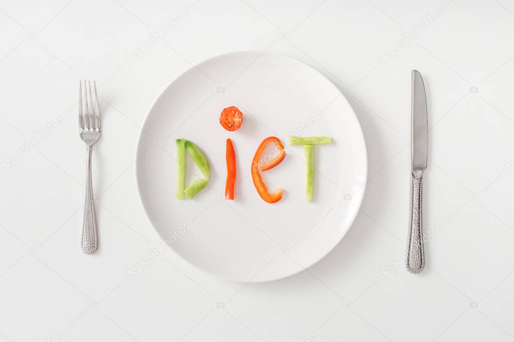 Top view of cutlery and diet lettering from vegetable slices on plate on white background