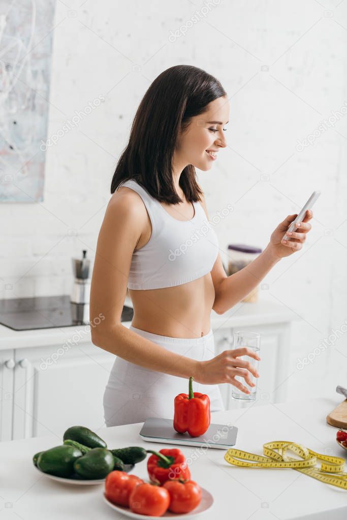 Side view of smiling sportswoman with smartphone and glass of water near vegetables and measuring tape on table, calorie counting diet