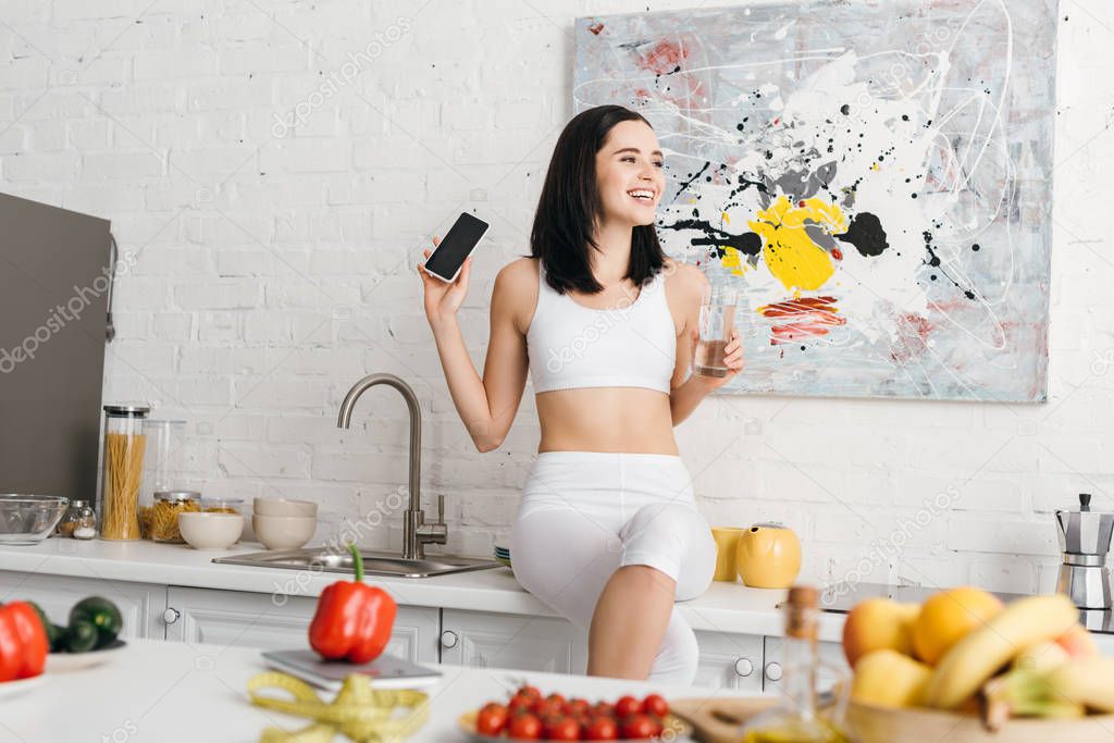 Selective focus of smiling sportswoman with glass of water and smartphone near vegetables, fruits and measuring tape on kitchen table, calorie counting diet