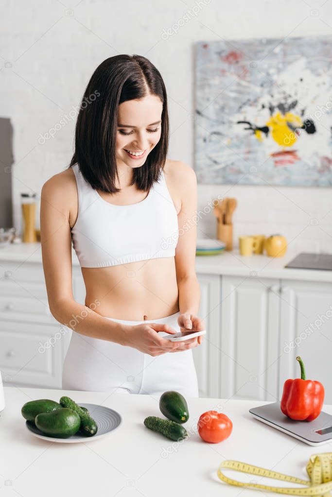 Beautiful smiling sportswoman using smartphone near vegetables, scales and measuring tape on table in kitchen, calorie counting diet