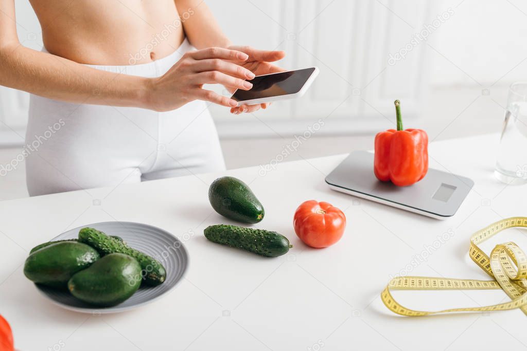 Cropped view of fit girl using smartphone near vegetables, measuring tape and scales on table, calorie counting diet