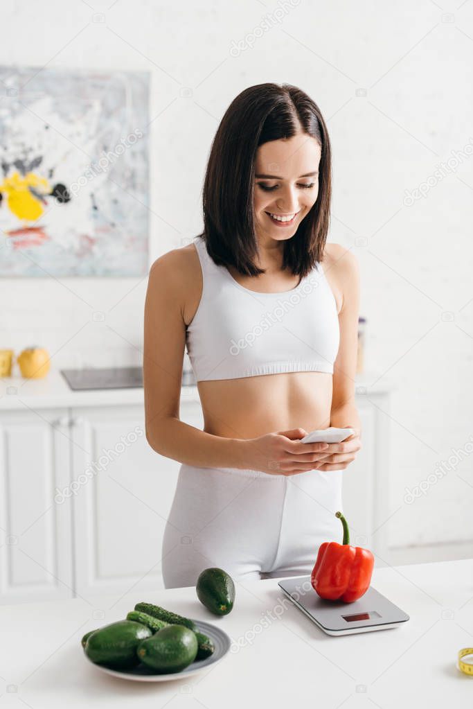 Smiling sportswoman using smartphone while counting calories of vegetables on scales on kitchen table, calorie counting diet