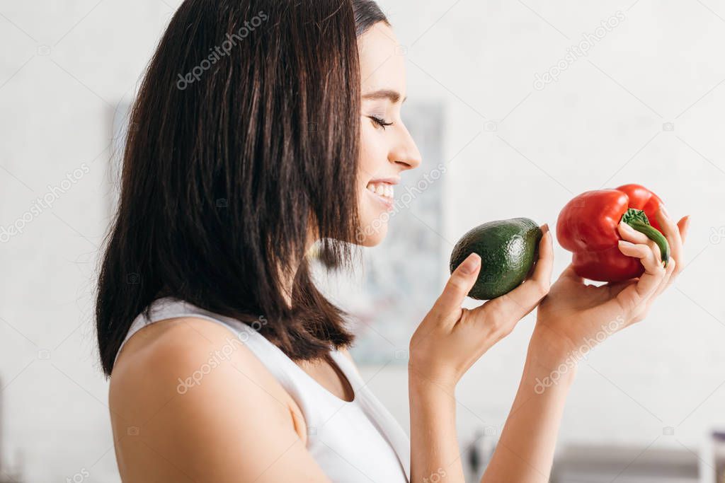 Side view of beautiful smiling woman holding ripe avocado and bell pepper in kitchen