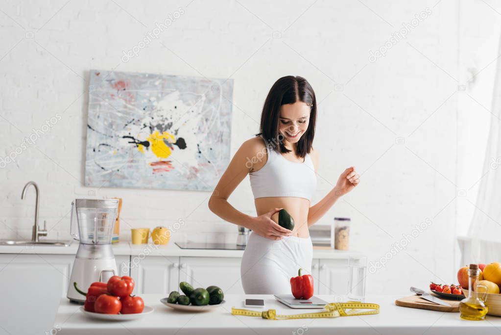 Smiling sportswoman holding avocado near fresh vegetables, scales and measuring tape on kitchen table, calorie counting diet