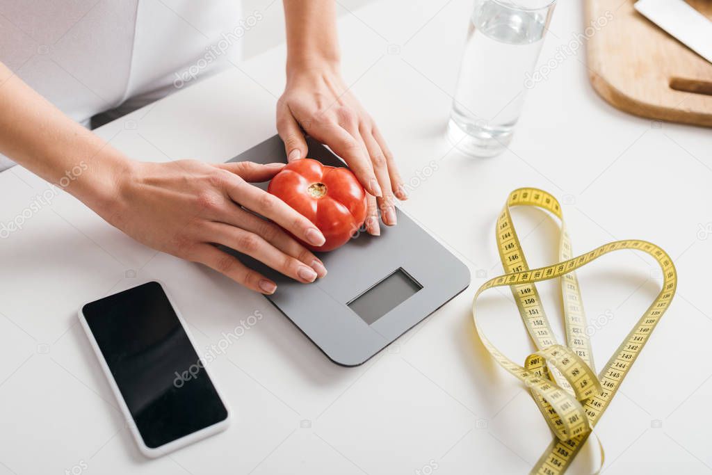 High angle view of woman putting tomato on scales near smartphone and measuring tape on kitchen table, calorie counting diet