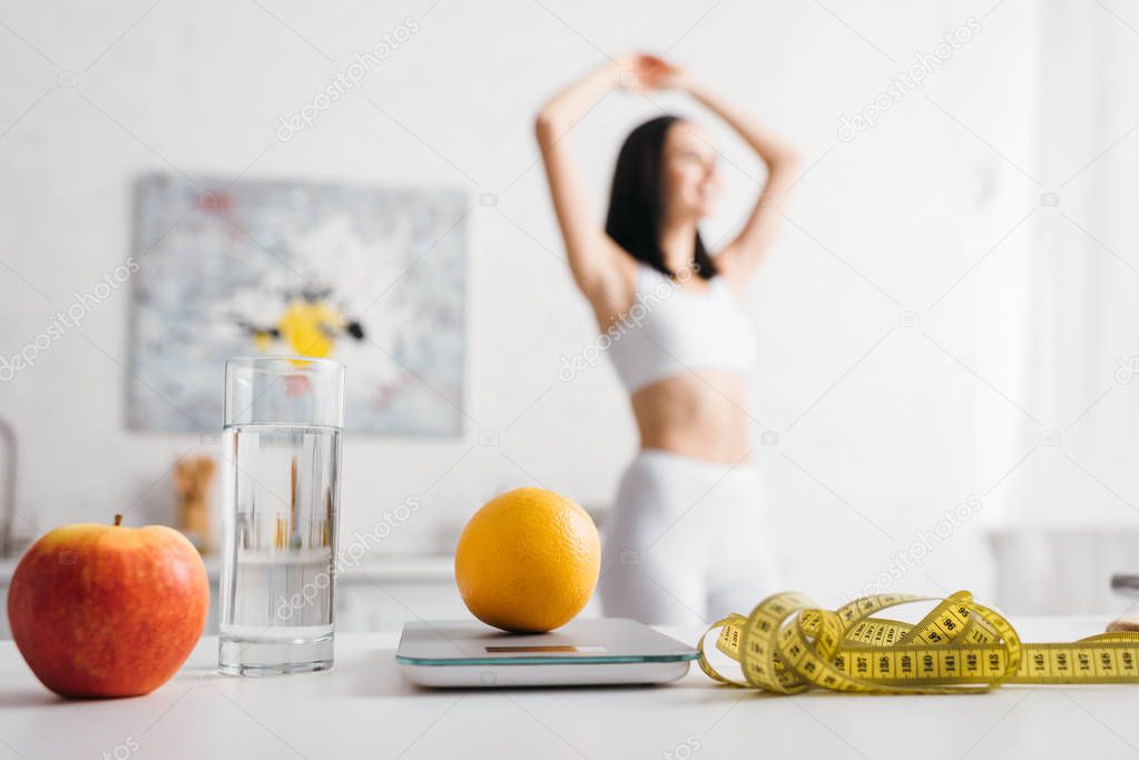 Selective focus of measuring tape, fruits with scales and glass of water on table and sportswoman in kitchen, calorie counting diet