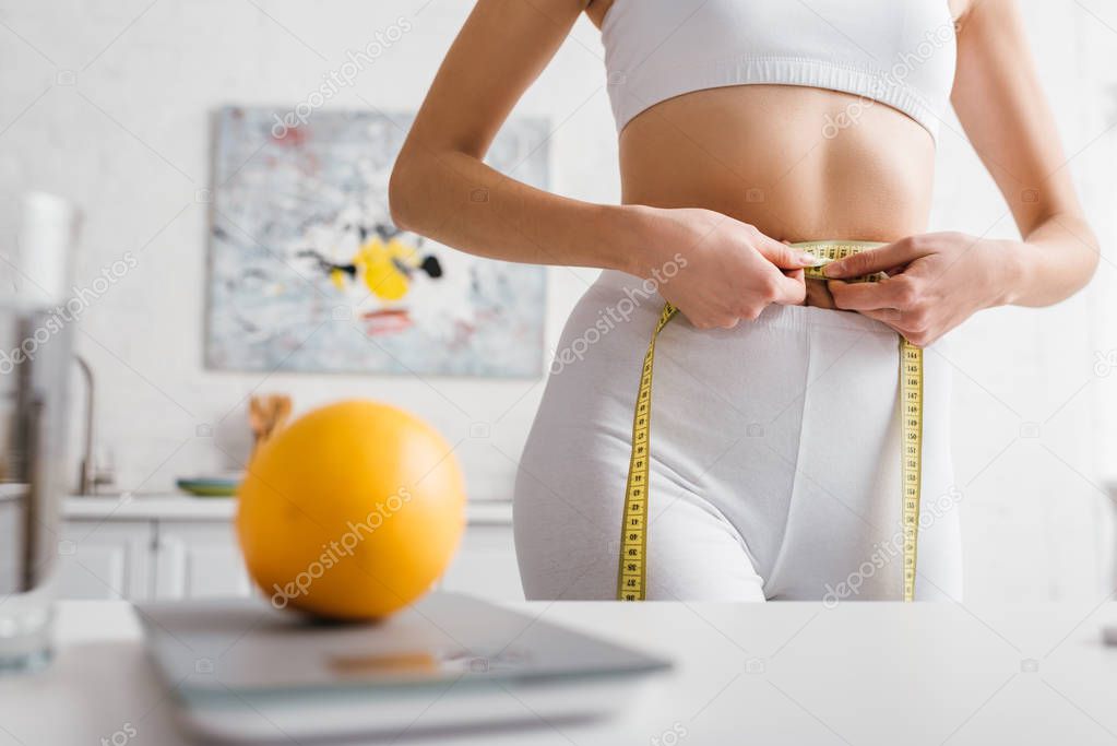 Selective focus of sportswoman measuring waist near orange on scales on kitchen table, calorie counting diet