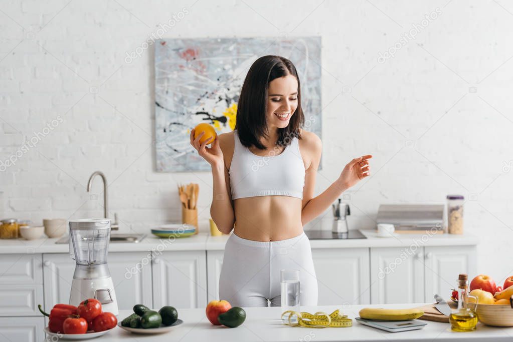 Smiling sportswoman weighing fruits near glass of water and measuring tape on kitchen table, calorie counting diet