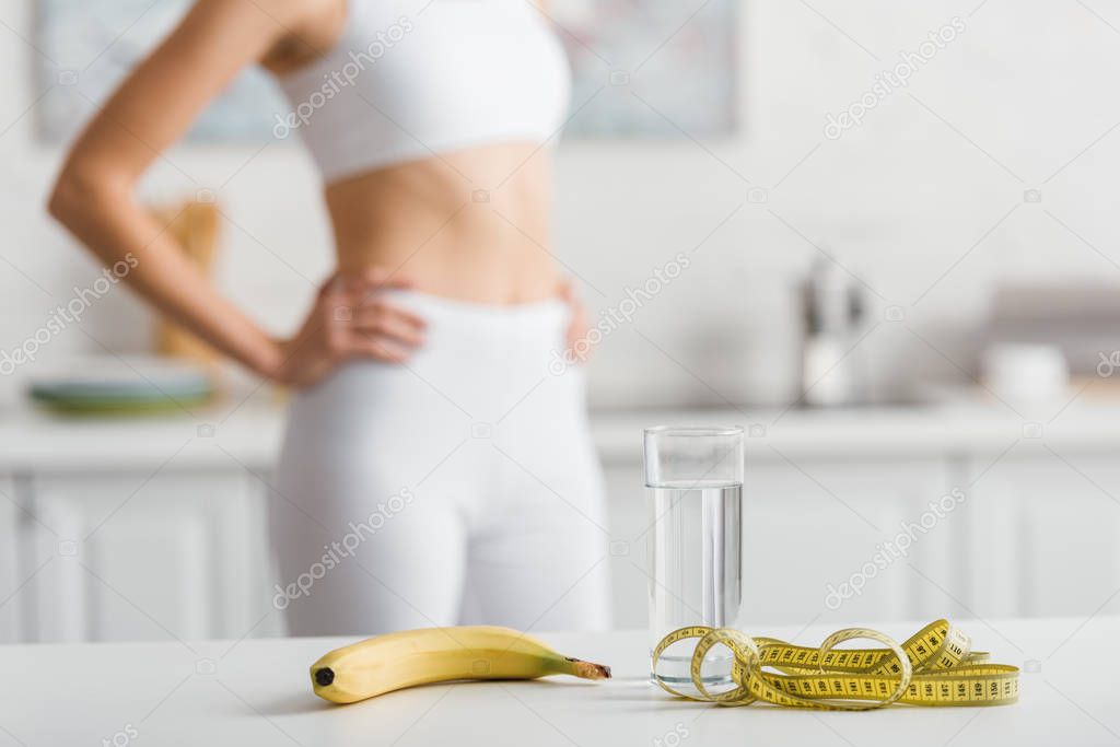 Selective focus of banana, glass of water and measuring tape on table near sportswoman in kitchen 