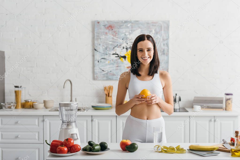 Smiling sportswoman holding orange near scales, measuring tape and vegetables on kitchen table, calorie counting diet