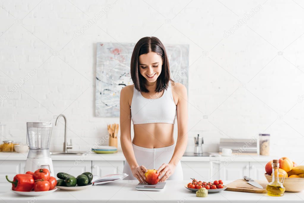 Beautiful smiling sportswoman weighing apple near vegetables, measuring tape and notebook on kitchen table, calorie counting diet