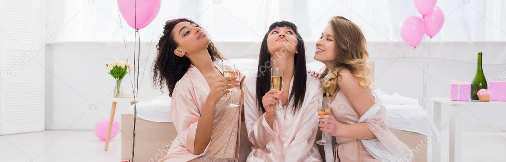 panoramic shot of positive multicultural girlfriends having fun and holding champagne glasses on pajama party with pink balloons