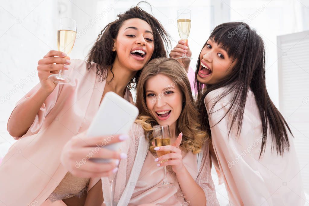 cheerful multicultural girls with glasses of champagne taking selfie on smartphone during pajama party