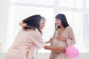 Pembe balonlu çok ırklı kızlar bebek partisinde hamile bir kadının karnına dokunuyor. 