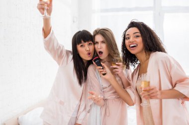 Heyecanlı çoklu etnik kızlar ellerinde şampanya ve pijama partisinde mikrofonla şarkı söylüyorlar.