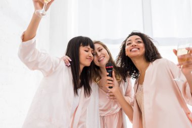 Güzel, mutlu, çok kültürlü kızlar ellerinde şampanya ve pijama partisinde mikrofonla şarkı söylüyorlar.