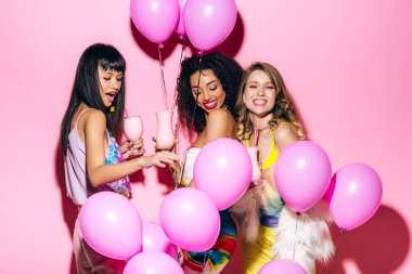 Neşeli, çok ırklı kızlar ellerinde balonlarla pembe milkshake tutuyorlar.