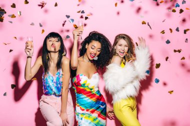 excited multicultural girls having fun with glasses of champagne on pink with confetti clipart