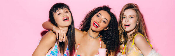 панорамный снимок счастливых модных мультиэтнических девушек, держащих стаканы с молочными коктейлями на розовом
