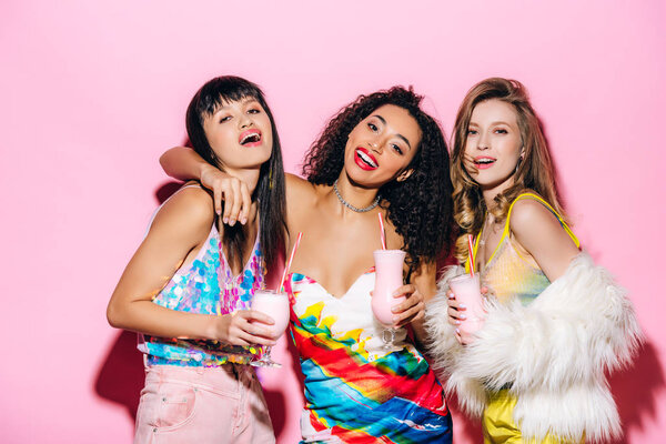 улыбающиеся стильные мультиэтнические девушки, держащие стаканы с молочными коктейлями на розовом

