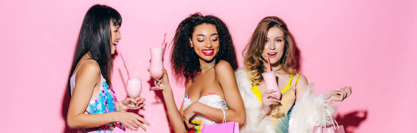 панорамный снимок веселых стильных мультиэтнических девушек, пьющих молочные коктейли на розовом
