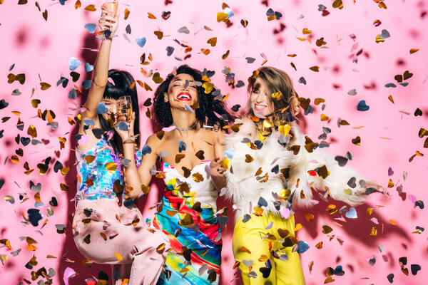 happy multicultural girls having fun with glasses of champagne on pink with confetti