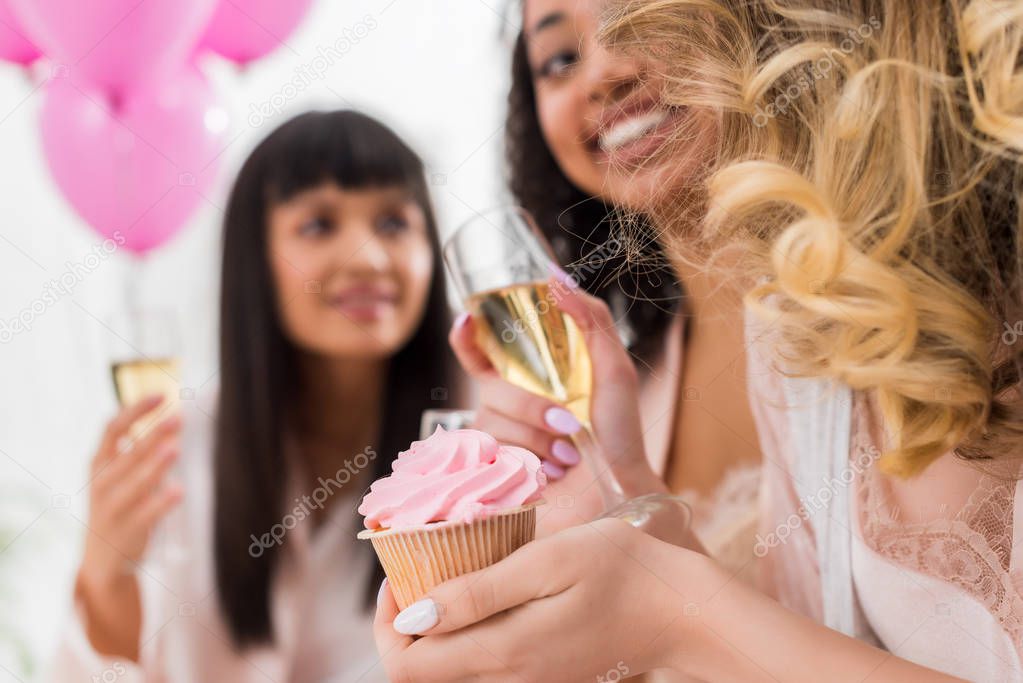 selective focus of happy multicultural girlfriends having fun with champagne and cupcake during bachelorette party 