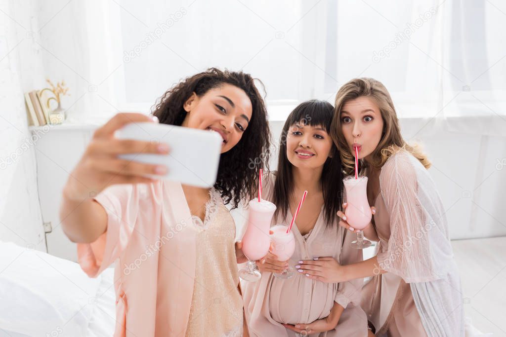 multiethnic girlfriends and happy pregnant woman drinking milkshakes while taking selfie on baby shower 