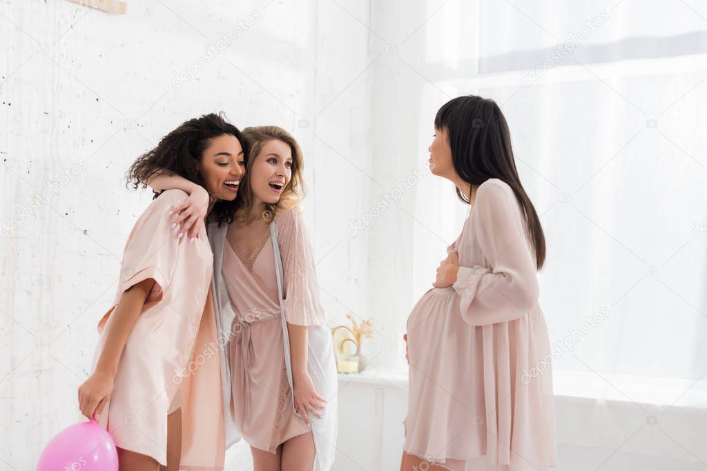 multicultural girlfriends with pink balloon looking at young pregnant woman on baby shower 