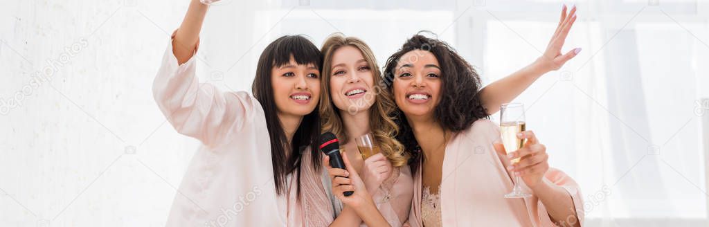 panoramic shot of attractive cheerful multicultural girls holding champagne and singing with microphone on bachelorette party
