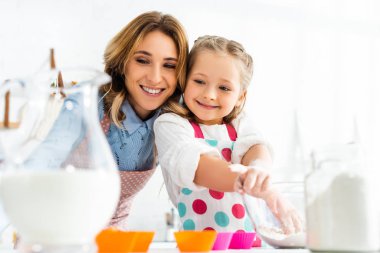 Selective focus of beautiful mother and daughter preparing cupcakes with ingredients including milk and flour in kitchen clipart