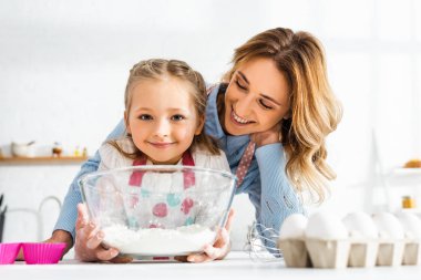 Selective focus of happy mom and cute daughter holding bowl with flour in kitchen clipart